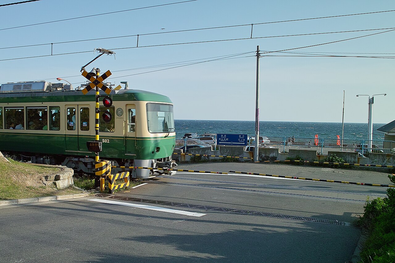 A green and cream Enoden train passing a crossing at Shichirigahama, with the sea behind it